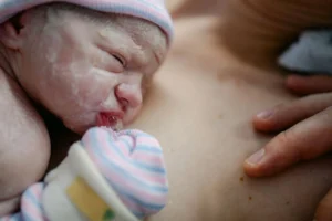 newborn wearing a striped hat resting on a caregiver’s bare chest