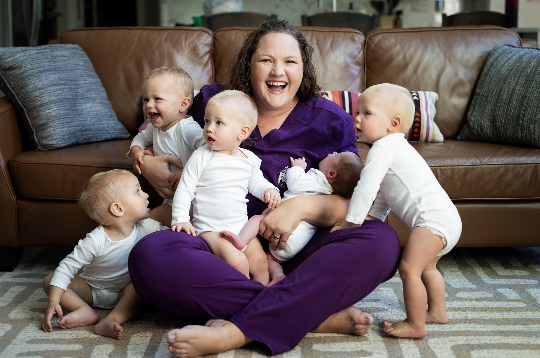 caregiver sitting on floor holding and surrounded by several infants in a cozy living room setting