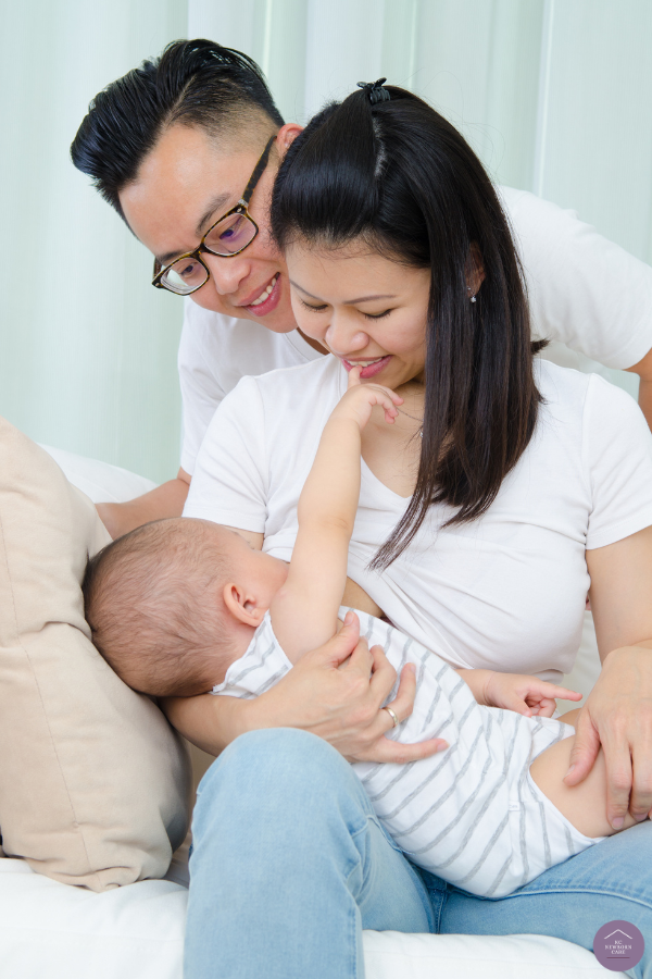 Mother nursing an infant while seated with partner leaning close beside her on a bed