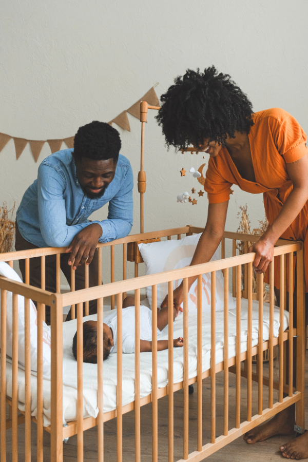 Parents leaning over a wooden crib to soothe an infant lying on a white mattress in a cozy nursery setting