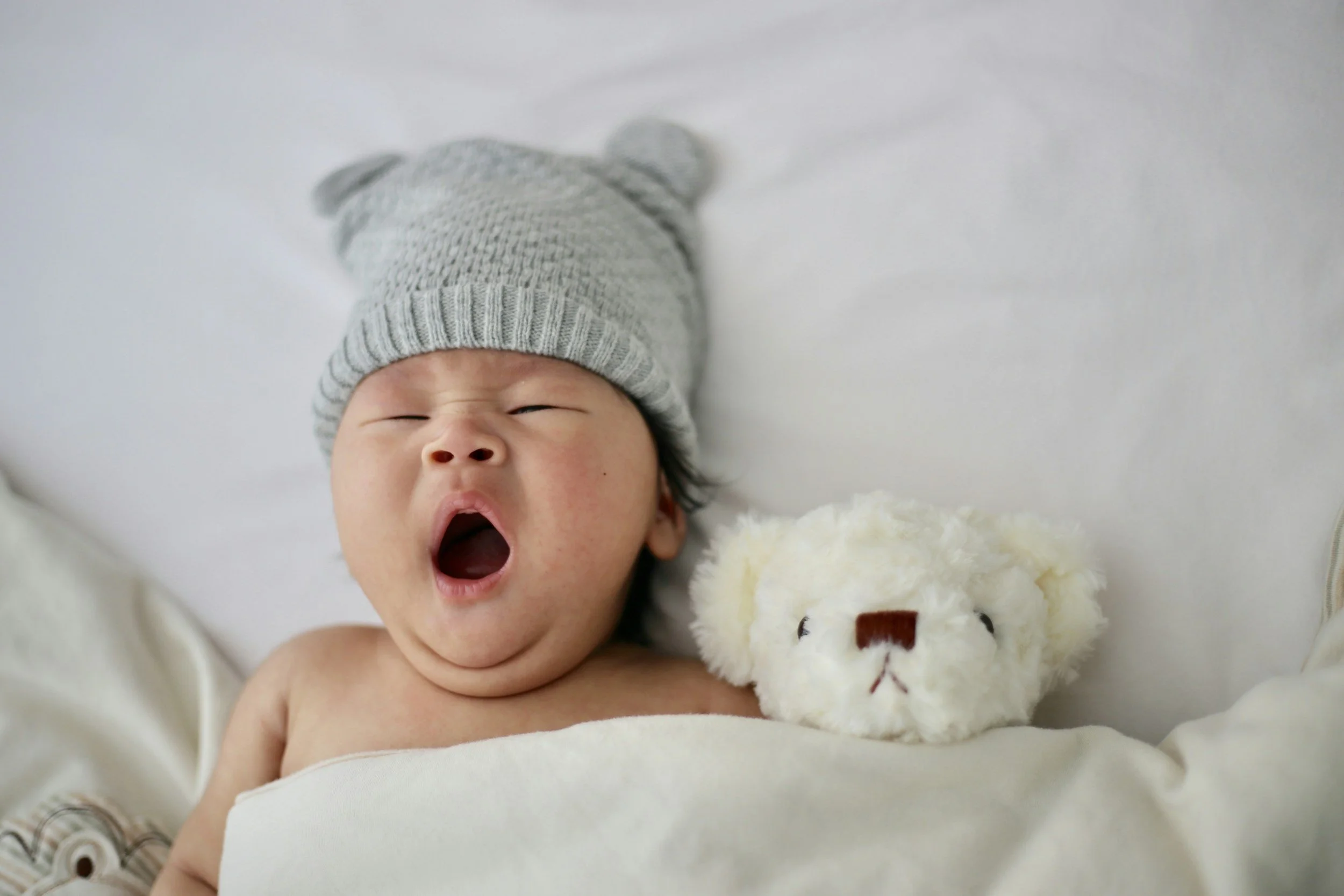 newborn lying under a blanket yawning beside a small white stuffed toy
