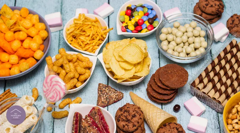Assorted sweet and savory postpartum snacks including chips, cookies, candy, and granola bars on a wooden table