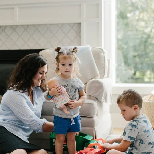 Caregiver sitting on floor with two young children playing with toys in a bright living room