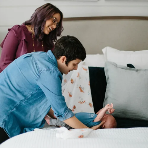 Parent gently changing a newborn on a bed while another adult watches nearby