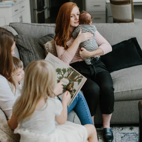 Caregiver holding a swaddled newborn while children look at a picture book on a living room couch